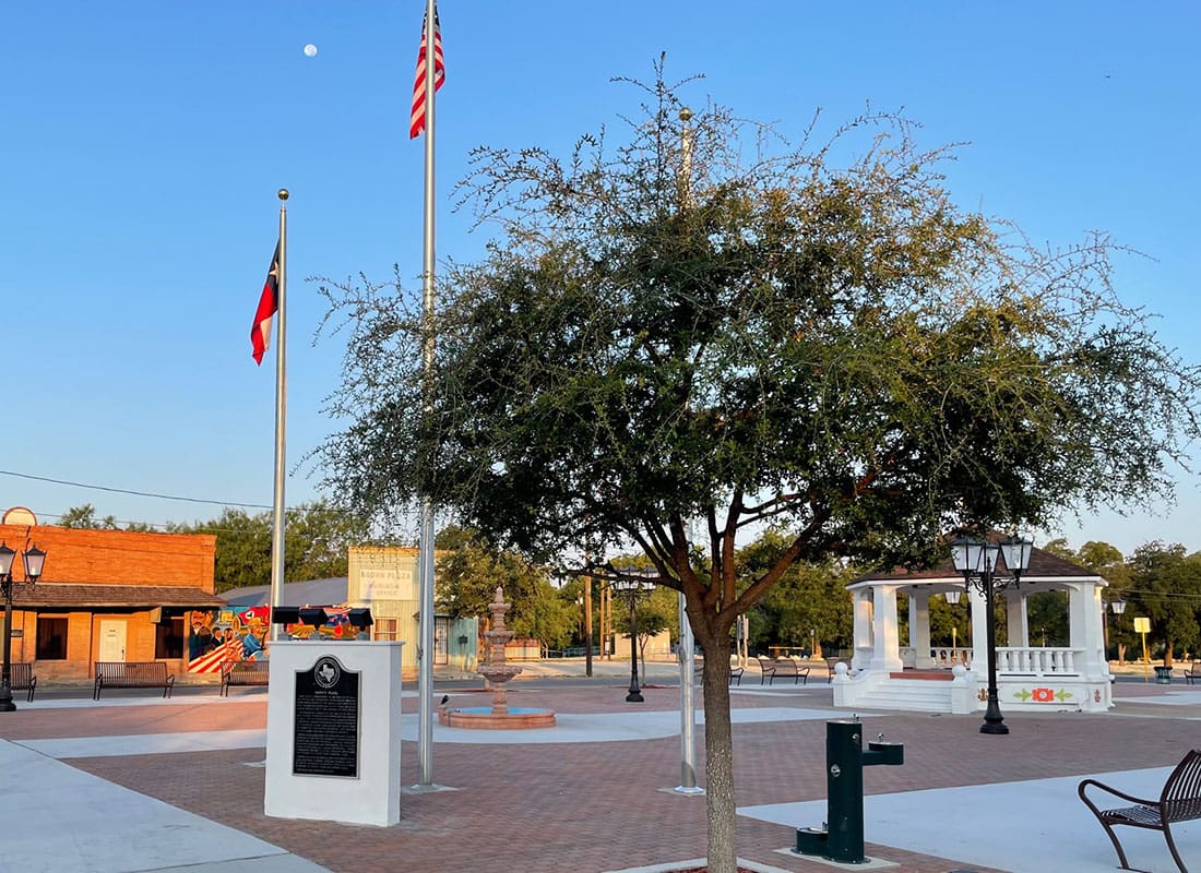 About Our Agency - View of a Tree in the Del Rio Town Square Next to a Historical Monument and Flags Against a Blue Sky